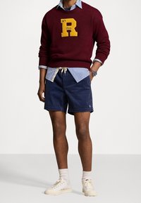 Bordeaux stickad tröja med gult "R"-logotyp, lager på lager över en blå och vit randig skjorta. Marinblå kordbyxor, beige dragsnöre, vita sneakers.