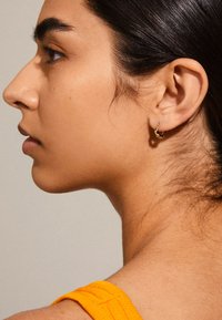 Gouden ringoorbel met een gedraaide ontwerp, voorzien van een gepolijste afwerking. Het model heeft donker haar en draagt een oranje getextureerde top.