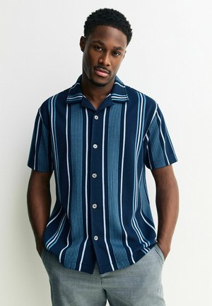 Bărbat în picioare, cu mâinile în buzunare, purtând o cămașă bleumarin cu mâneci scurte, cu dungi verticale albastre deschis și albe, și pantaloni gri.