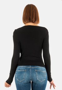 Donna con capelli corti castani che indossa un top a maniche lunghe nero e jeans blu, vista di spalle su uno sfondo bianco semplice.