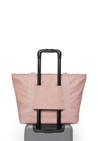 Hellrosa Tragetasche mit dicken, flachen Riemen, entworfen, um über einen Koffergriff zu passen, die auf einem grauen Hartschalenkoffer ruht.