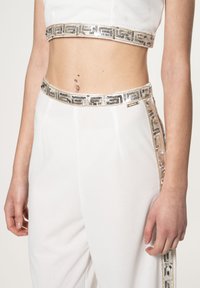 Top corto bianco con fascetta metallica decorata, abbinato a pantaloni bianchi con una striscia laterale in paillettes e una texture di tessuto liscia.