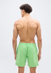 Άνδρας χωρίς μπλούζα με κοντά, σγουρά μαλλιά, που στέκεται με την πλάτη γυρισμένη, φορώντας φαρδύ σορτς με ελαστική μέση φωτεινού πράσινου χρώματος, πάνω σε απλό λευκό φόντο.