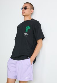 Sort bomulds T-shirt med et grønt kaktus grafik og hvid tekst, parret med lyse lilla Nike shorts. Modellen bærer sølv solbriller.