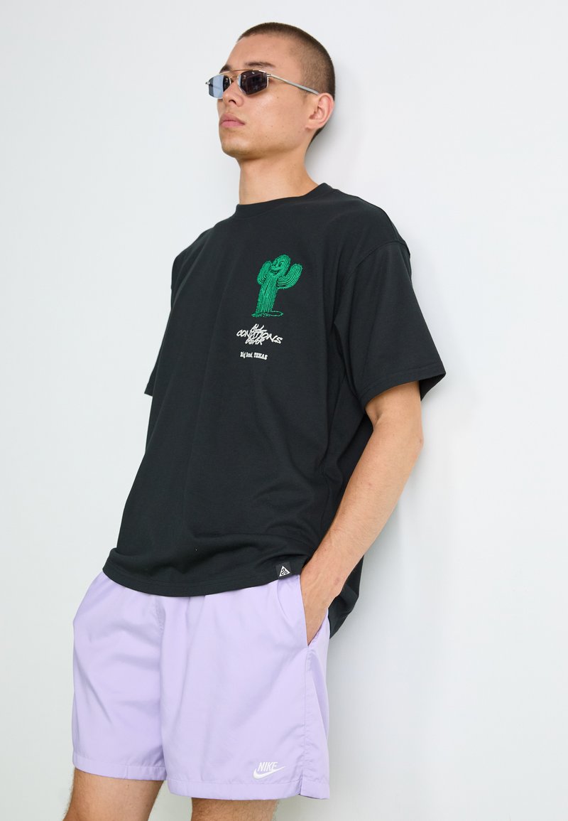T-shirt en coton noir avec un graphique de cactus vert et du texte blanc, associé à un short Nike violet clair. Le modèle porte des lunettes de soleil argentées.