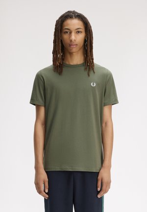 Giovane con lunghi dreadlocks che indossa una maglietta verde oliva e pantaloni blu scuro, in piedi su uno sfondo bianco semplice.