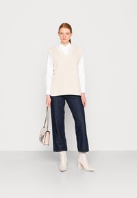 Gilet a coste beige con scollatura a V, indossato sopra una camicia bianca. Abbinato a pantaloni larghi in denim scuro e stivaletti beige. Tiene una borsa leggera.