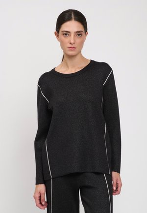 Donna con i capelli scuri raccolti in una coda bassa, indossa un top nero a maniche lunghe e pantaloni coordinati con dettagli di cuciture bianche su sfondo uniforme.
