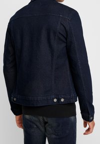 Chaqueta de denim azul oscuro con cierre frontal de botones, acentos de hardware metálico y un diseño estructurado. Presenta una banda en la cintura con broches a presión.