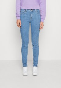 Jean skinny bleu clair en denim, avec cinq poches et une taille standard. Associé à des baskets blanches et un sweat-shirt lavande.