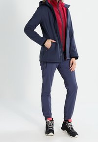 Person wearing a navy jacket, red fleece zip-up, navy pants, pink socks, and black hiking boots, standing with one hand in pocket.