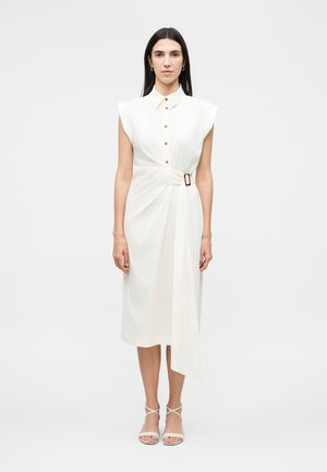 Kvinde med langt mørkt hår iført en ærmeløs hvid kjole med knapper og et bælte i taljen, stående mod en ensfarvet lys baggrund.