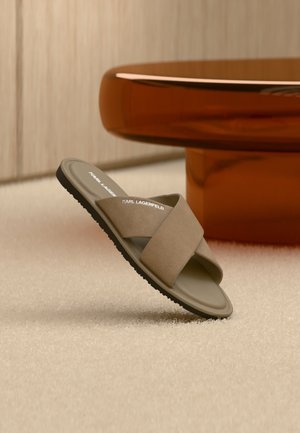 Une seule sandale beige à glissière avec des brides croisées et le logo "Karl Lagerfeld", posée sur un tapis clair près d'un objet circulaire marron.