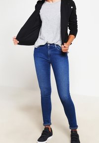 Jeans skinny blu scuro in denim, con vita alta, due tasche frontali e orli arrotolati. Abbinati a una t-shirt grigia e a un giubbotto nero.