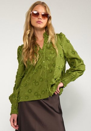 Blonde Frau mit großen Sonnenbrillen, einer strukturierten olivgrünen Bluse mit Knöpfen und floralen Akzenten sowie einem dunkelbraunen Rock, die mit der Hand in der Hüfte posiert.