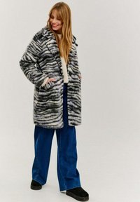 Kabát z umelého kožušiny s čierno-bielym vlnitým vzorom, veľké klopy, bočné vrecká, v kombinácii s modrými širokými nohavicami a čiernymi topánkami.