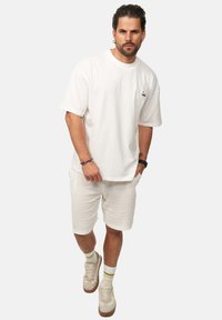 Weißes Baumwoll-T-Shirt mit kurzen Ärmeln und Logo auf der Brust, kombiniert mit weißen strukturierten Shorts und beigen Sneakers mit gelben und grünen Akzenten.