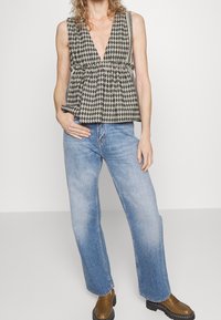 Top peplum a cuadros en beige y negro, con cuello en V profundo, combinado con jeans de pierna ancha en azul claro y mocasines marrones, de pie contra un fondo neutro.