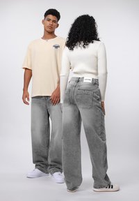Camiseta beige de gran tamaño, vaqueros grises de pierna ancha, suéter texturizado blanco de canalé; el modelo posa de espaldas mostrando los detalles del atuendo. Zapatillas blancas visibles.