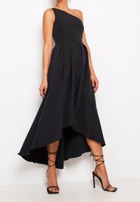 Robe noire à une épaule avec un corsage ajusté et une jupe asymétrique plissée, présentant une texture lisse. Portée avec des talons noirs à brides.