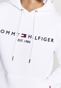 Felpa bianca con cappuccio, dotata di coulisse con punte metalliche, logo ricamato "TOMMY HILFIGER" e "EST. 1985" in blu e rosso.