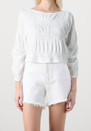 Kobieta ubrana w białą, długorękawową bluzkę z koronkowymi detalami oraz białe, przetarte szorty z wysokim stanem na jednolitym tle.