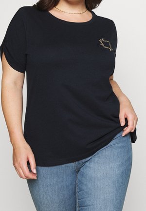 Frau trägt ein schwarzes kurzärmliges Shirt mit einer kleinen goldenen Kettenhalskette und blauen Jeans, hält den Saum des Shirts auf einer Seite fest.