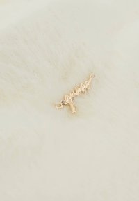 Arany színű emblémacharm, amelyen a "guess" felirat olvasható, textúrázott fehér szőrmével háttérben, sima ívekkel és egy kis díszkővel.