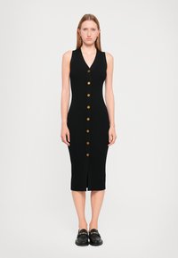 Robe midi noire côtelée avec un décolleté en V, ornée de boutons dorés sur le devant et d'une fente à l'avant. Portée avec des mocassins noirs épais.