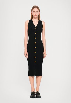 Zwart geribbelde midi-jurk met een V-hals, voorzien van gouden knopen langs de voorkant en een split aan de voorkant. Gecombineerd met chunky zwarte loafers.