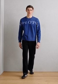 Sudadera de algodón azul con el logo "Marc O'Polo" en blanco, combinada con pantalones negros y zapatillas negras; diseño sencillo, ajuste relajado y mangas largas.