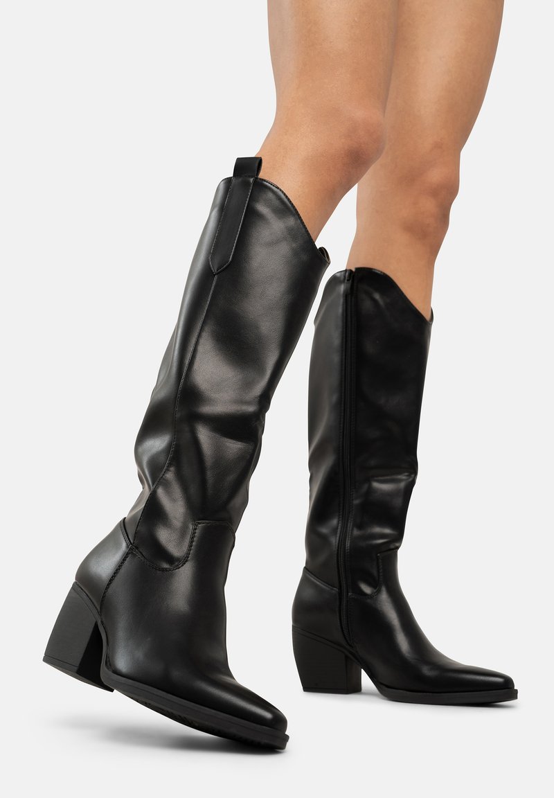 Botas de cuero negro hasta la rodilla con punta afilada, detalle recortado, cremallera trasera y tacón apilado, con una textura suave y un diseño elegante.