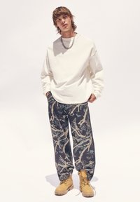 Chemise blanche à manches longues, pantalon ample à motifs sombres avec un design vert et beige, et bottes jaunes. Comprend un collier en chaîne.