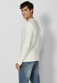 Lichtgrijs longsleeve shirt met verticale ribbelstructuur, ronde hals en aansluitende mouwen, gecombineerd met blauwe spijkerbroek.