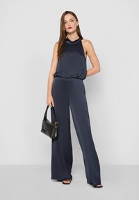 Námornícky modrý overal s bezrukávovým dizajnom a prehodeným výstrihom. Má nohavice s širokými nohavicami a hladký materiál. Doplněný čiernou kabelkou.