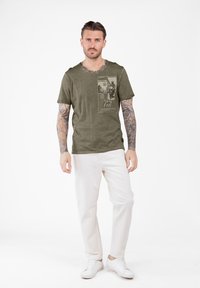 Camiseta de manga corta verde olivo con un estampado gráfico en la parte frontal, corte holgado, textura de tela suave, combinada con pantalones blancos y zapatillas.
