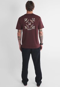 Homme tatoué portant un t-shirt bordeaux avec un graphisme floral et le texte "OXBOW", un pantalon noir, se tenant de dos sur un fond blanc.