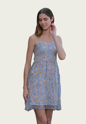 Jeune femme portant une robe sans manches à imprimé floral avec de fines bretelles, touchant son cou et souriant doucement sur un fond clair.