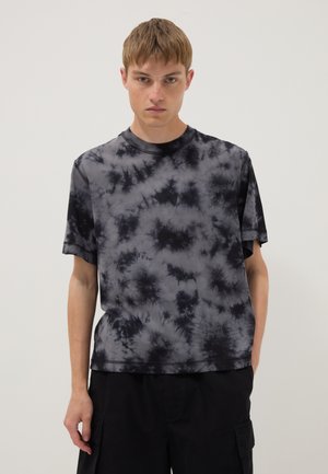 Giovane uomo con una t-shirt a maniche corte tie-dye nera e grigia e pantaloni neri, in piedi davanti a uno sfondo chiaro e uniforme.