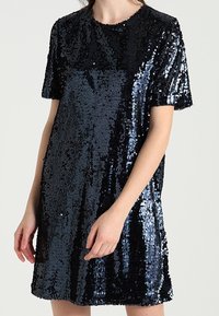 Kvinna som bär en kortärmad svart paljettklänning med rund halsringning mot en enfärgad bakgrund.