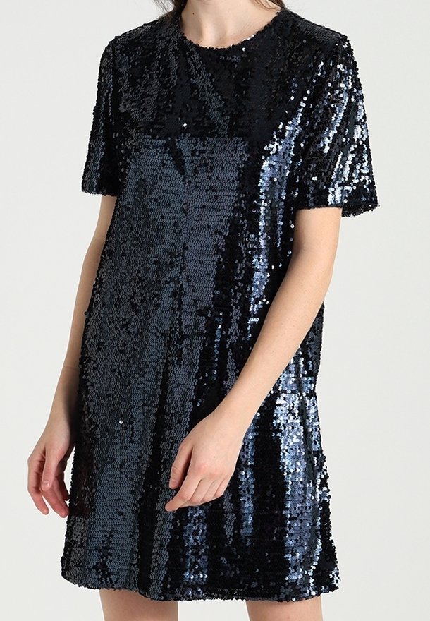 Kvinna som bär en kortärmad svart paljettklänning med rund halsringning mot en enfärgad bakgrund.