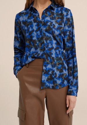 Vrouw draagt een blauw-zwarte bloemenblouse met lange mouwen, ingestopt in hooggetailleerde bruine leren broek met zijzakken.