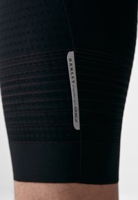 Șorturi sportive negre cu puncte texturate, accente dungi în roșu și un eticheta argintie cu inscripția "OAKLEY PWRD BY Q365" pe lateral.