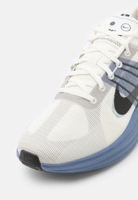Gros plan d'une chaussure de course blanche Nike avec semelle bleue, swoosh noir et lacets blancs sur un fond clair uni.