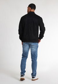 Jachetă neagră din suede cu guler, asortată cu blugi denim albaștri și adidași albi, surprinsă din spate pe un fundal simplu.