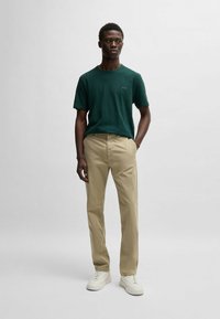 Camiseta verde de manga corta con logo, pantalones rectos beige y zapatillas blancas, con un fondo claro y sencillo.