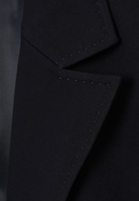 Gros plan sur le revers d'une veste noire montrant les détails de la couture fine et la texture lisse du tissu.