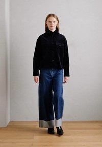 Veste en velours côtelé noire avec fermeture à boutons et deux poches poitrine, associée à un jean bleu à jambes larges avec des ourlets argentés. Simples bottines noires à la cheville.