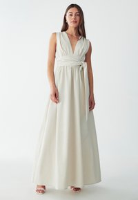 Robe maxi sans manches de couleur beige clair avec un décolleté en V profond, une taille ajustée et un nœud sur le côté. Fabriquée dans un tissu doux et texturé. Silhouette lisse et fluide.