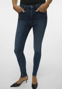 Jeans skinny blu scuro in denim, dotati di tasche frontali e vita alta. Il tessuto ha una leggera lucentezza e una consistenza liscia.